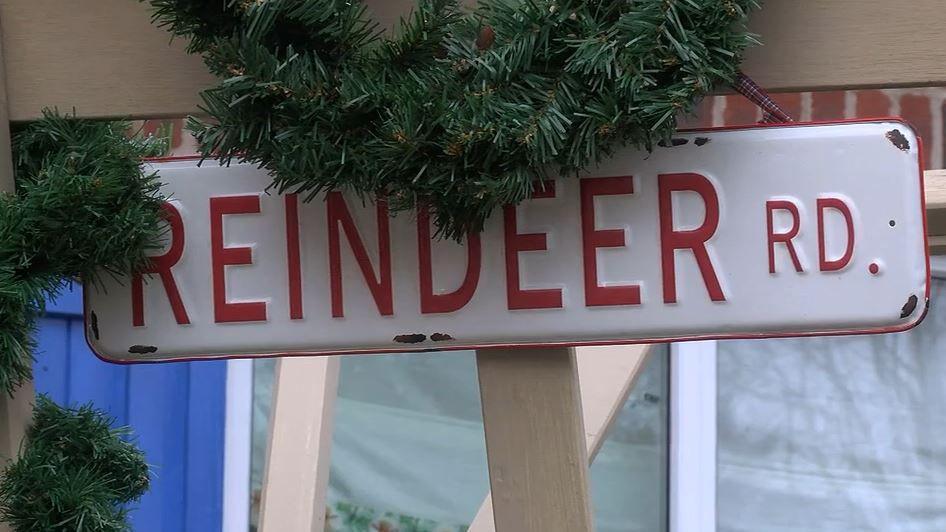 A white and red sign reading 'Reindeer RD' on a piece of wood. A Christmas wreath is hung above it. 