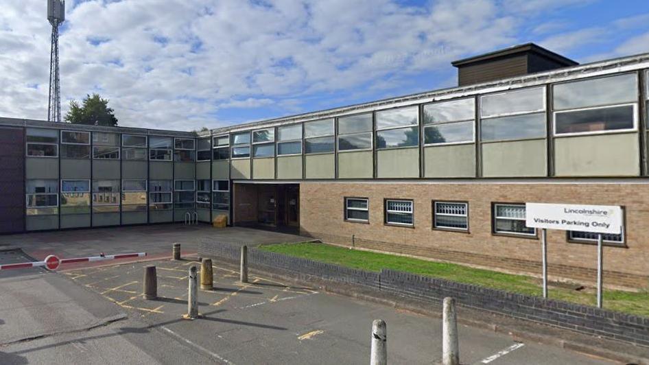 A 1960s two-storey office building, dominated by glass windows and green panels. In the foreground, a car park with a red and white barrier. A white sign reads: Lincolnshire County Council, visitors parking only.