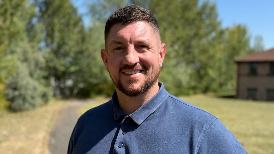 Colin Burns, who has brown gelled hair and a brown beared, is wearing a dark blue button-down T-shirt. He is smiling in front the camera as he stands on a concrete path. In the background, there are tall trees and faded grass. To his right, there is an office building which has a red roof and beige coloured bricks.