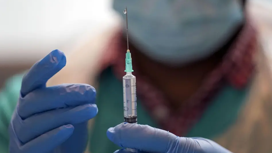 A medic is wearing gloves and holds a needle close to the camera while their face and mask is blurred behind it