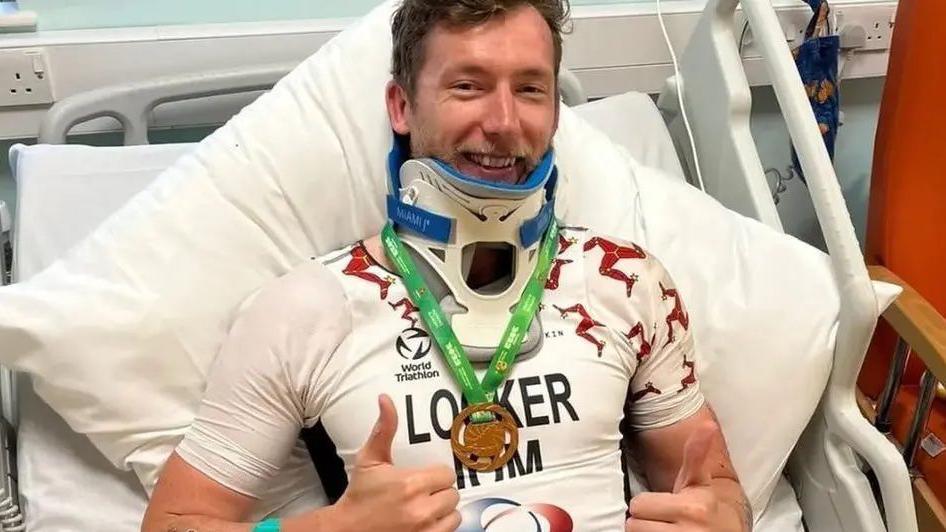Matt Looker wears a white and red lycra top in a hospital bed with a pillow propping him up. He wears a neck brace, and a bronze medal with a green ribbon has been hung around his neck. He smiles and does two thumbs ups.