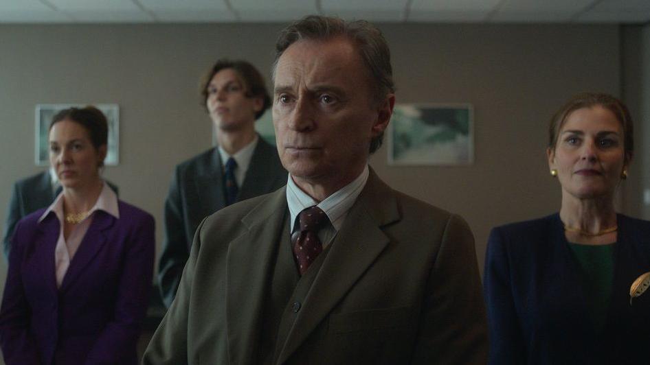 Robert Carlyle as Sam Hagen. He is stood in a suit with other actors playing members of the council in a drab office room