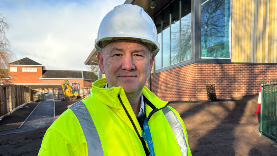 A smiling man with a yellow high-visibility jacked and white construction-workers helmet standing in front of a building with large windows, wood panelling and red brick walls