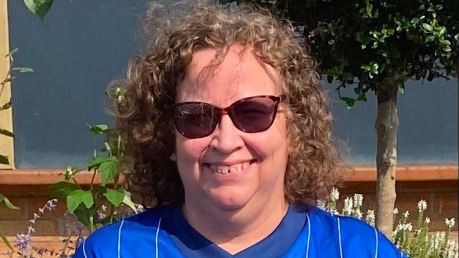 A woman smiles at the camera while sitting outside. She has shoulder length, brown curly hair and she is wearing sunglasses along with a blue football top.