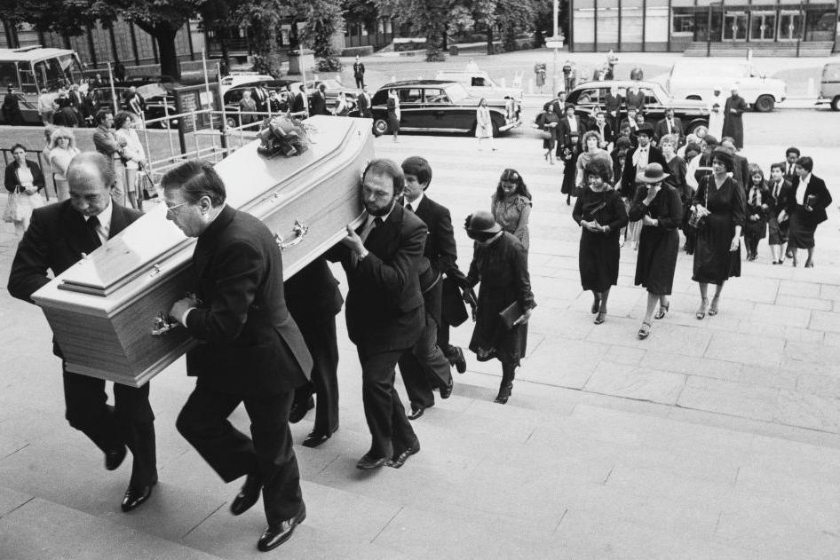 Pall bearers carry a coffin at the funeral of Dr Amal Dharry up the steps of Coventry Cathedral, with mourners following on behind with funeral cars parked in the background