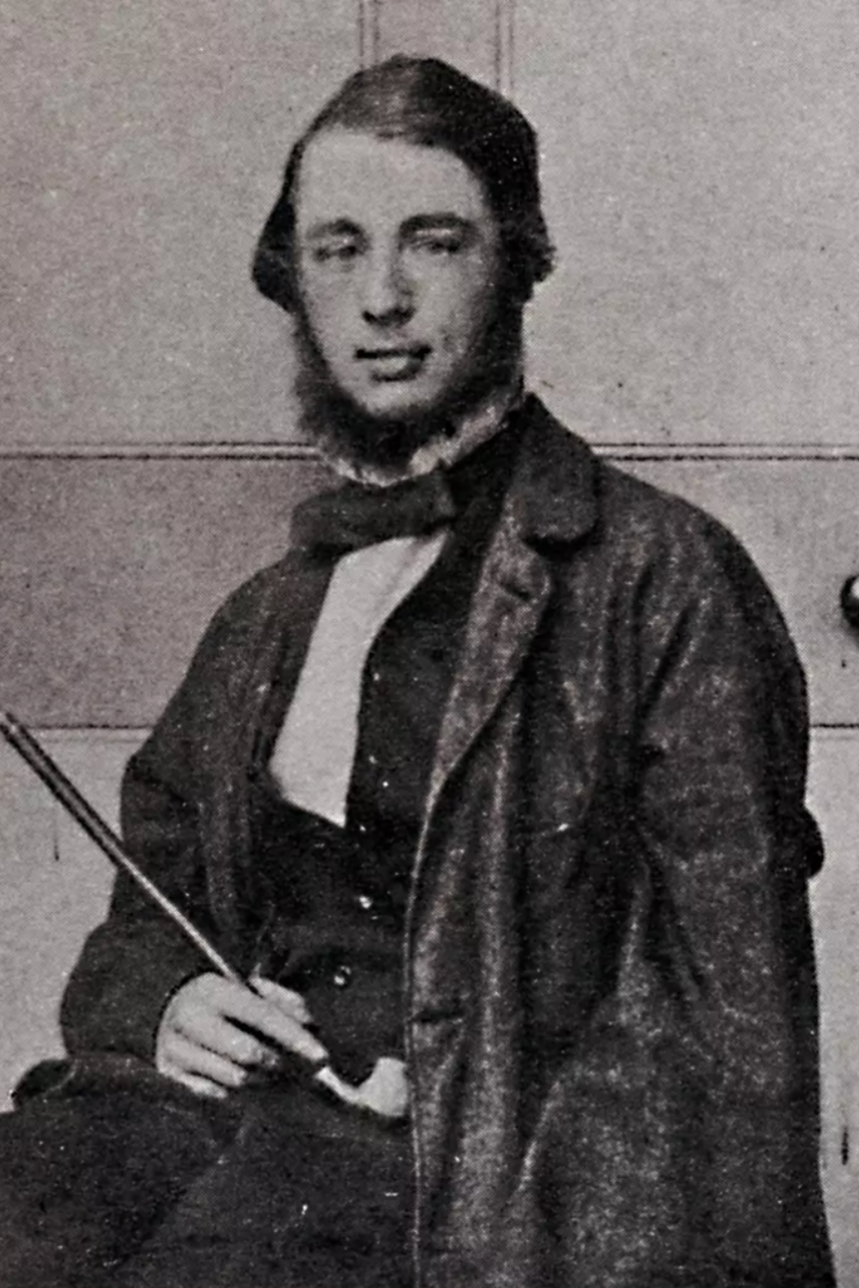A bearded man wearing a top coat, ruffle-neck shirt and bow-tie looks towards the camera. He is carrying a long-handled pipe
