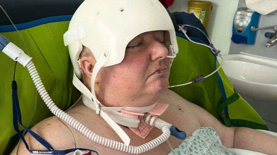 Georgina Price in a hospital bed post surgery with a tracheotomy - wearing a white helmet used to protect the surgical site