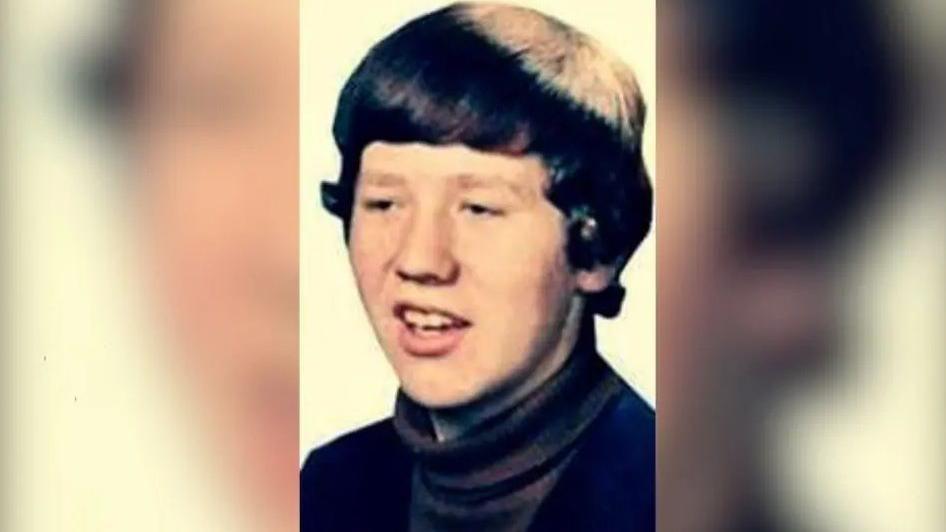 Columba McVeigh is looking left of the shot. It is a headshot of him and he has his mouth open. He has dark hair and is wearing a dark turtleneck jumper.