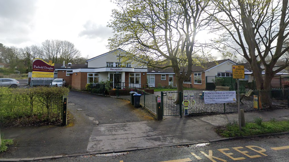 A Google Streetview of the exterior of Parbold Douglas Church of England Academy. It is a single-storey building with a Christian cross on the outside. There is a sign outside the school in the car park.