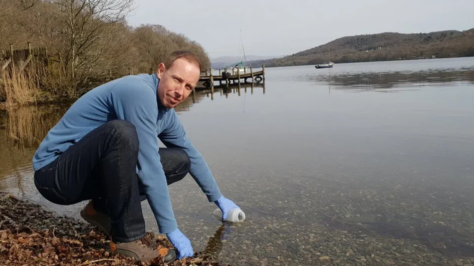 Man in blue top holding bottle to a lake while looking at the camera