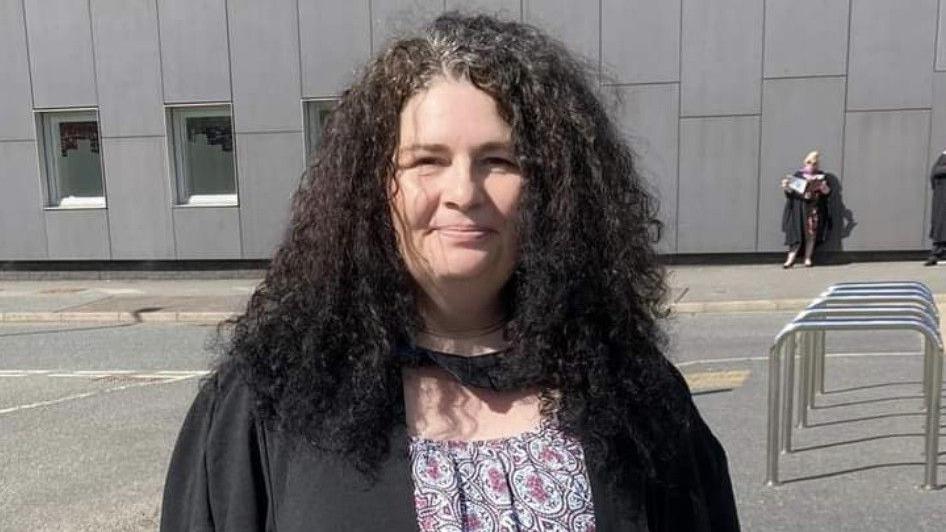 A middle-aged white woman standing outside a large grey building. She is wearing a graduation gown and has black hair. She is smiling at the camera.
