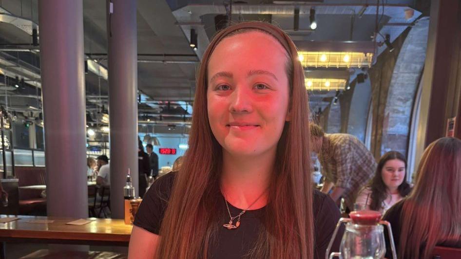 Rebecca is sitting in a restaurant. She is smiling at the camera wearing a black T-shirt