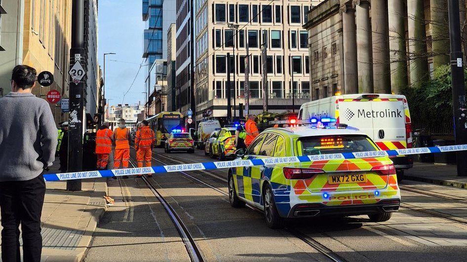 Metrolink workers in orange hi vis gather on a tram track in Manchester city centre. In the background, a tram is just visible, in the foreground there are several police cars with their lights on parked up on the tram track, which is cordoned off with police tape