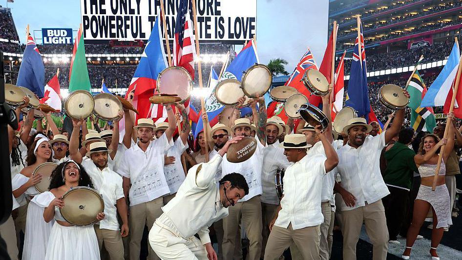 Bad Bunny spikes a football during his Super Bowl half-time show in 2026, surrounded by dancers waving flags and tambourines