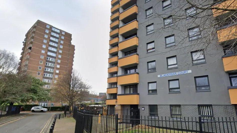 The apartment at Black Friar Court in Salford from where William Waheed Lyall fell. It is a grey building with yellow balconies. It is surrounded by metal fencing and bollards. Another apartment building is on the left of the picture with a few trees lining the road. 