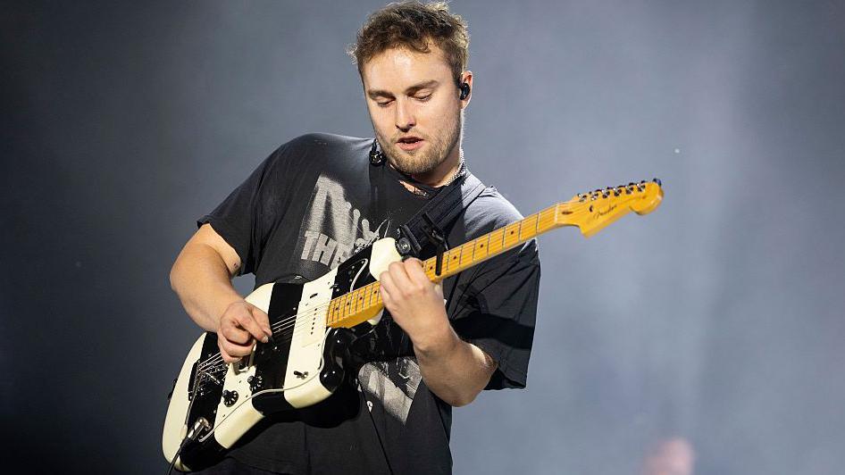 Sam Fender on stage with smoke behind him. He is playing a white and black guitar and wears a black t-shirt.