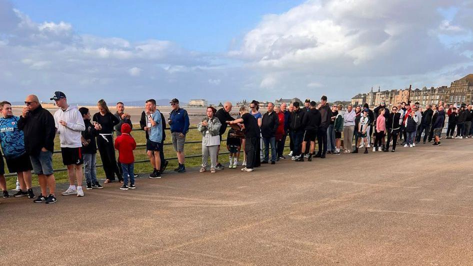 A queue of people along a beach promenade. In the background there are some buildings on the right hand side, and there is a cloudy blue sky above.
