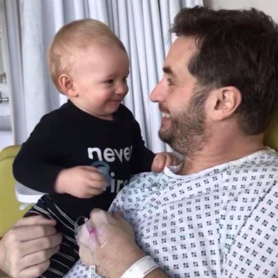 Gavin is wearing a hospital gown, holding Fletcher. They're both looking at each other and smiling.