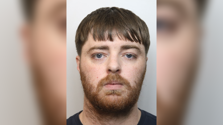 Callum Harvey-Mackenzie looking at the camera in a police mugshot. He has a long fringe and a full beard which is ginger.