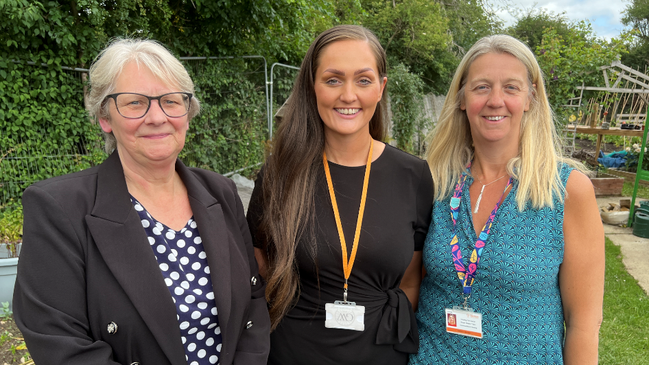 Cllr Howarth (L), Chelsea Eddolls and and Becky Davidson all smile at the camera. Cllr Howarth is wearing a black jacket and a navy and white spotty top. Chelsea wears a black dress with an orange lanyard and Becky wears a blue dress with a multicoloured lanyard. They are standing in a garden with greenery around them.