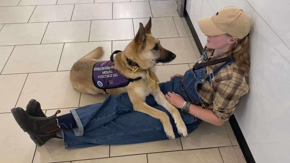 A young woman sits against a wall wearing dungarees and a cap. A German Shepherd dog lies on her lap.