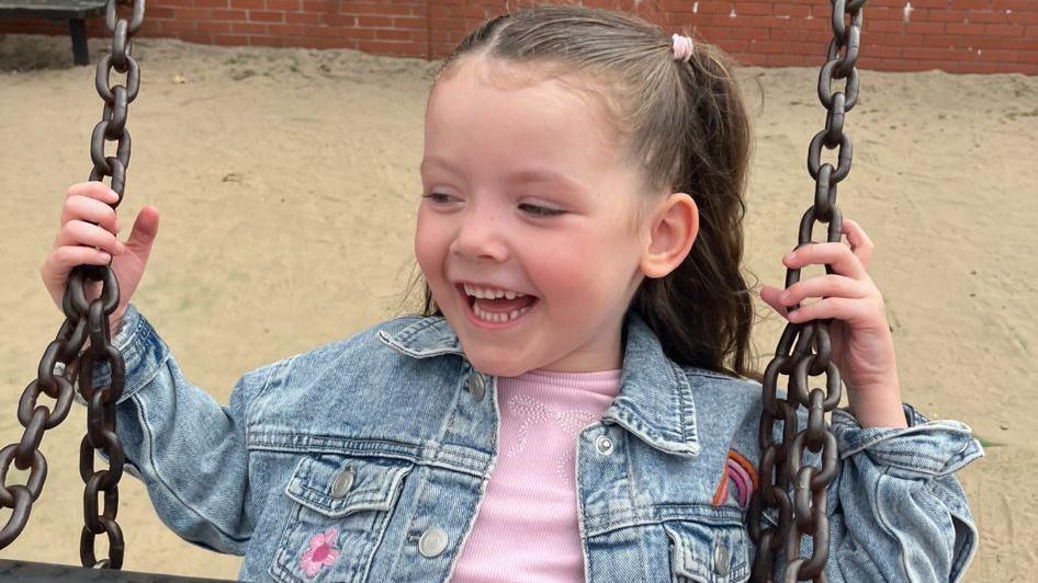A photo of Niamh on a swing in a playpark. She has her hair in pigtails and is wearing a blue denim jacket over a pink shirt