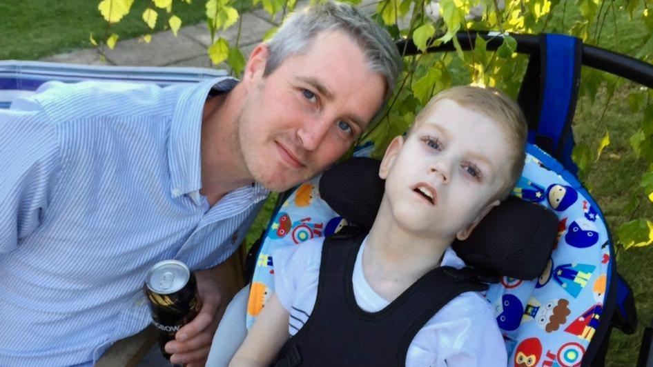 Shaun, wearing a blue shirt and holding a beer, smiles at the camera, next to Toby