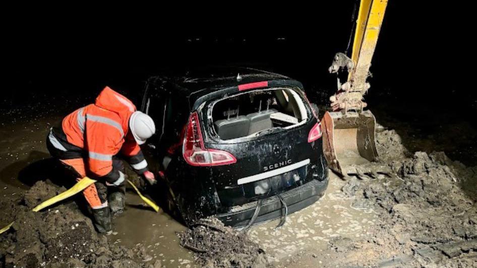 A black car stuck in watery mud with a worker in orange high vis leaning down and working on retrieving the car by attaching rope to it.