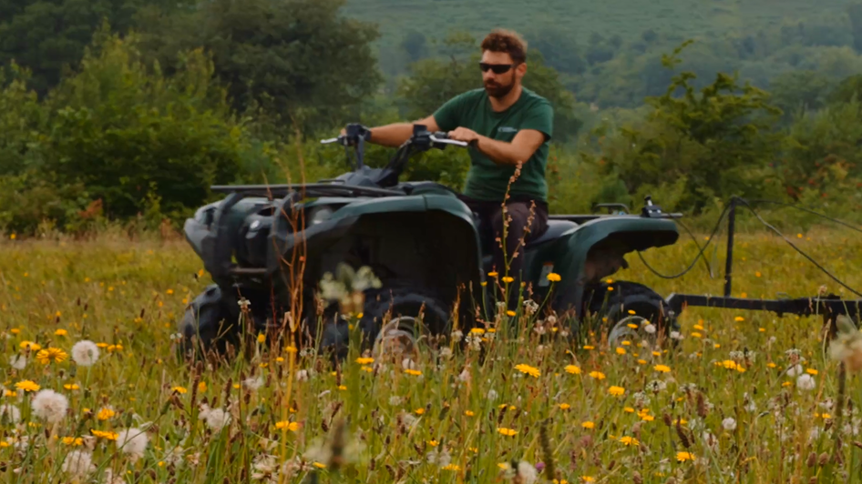 A field lush with yellow, white and pink wildflowers. A man on a quadbike is towing a red machine behind him.