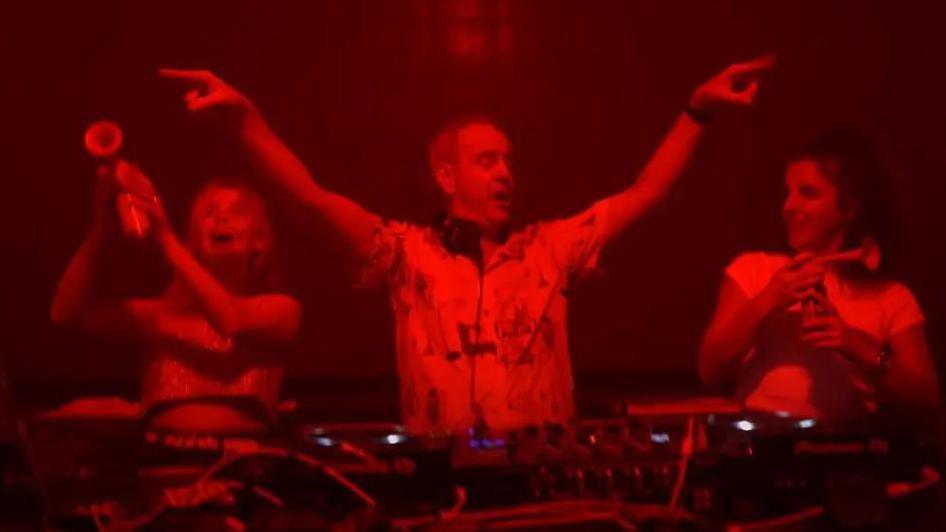 Saoirse-Monica Jackson and Jamie-Lee O'Donnell stand on stage behind a DJ deck with DJ Fatboy Slim holding air horns during his gig in Londonderry. The DJ stands in between the two women with his hands in the air. The image is dark with a red light hue over the top.