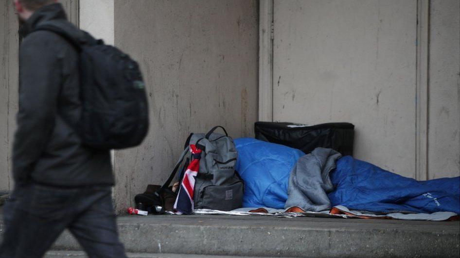 A person, whose face cannot be seen, lies in a blue sleeping bag in a doorway surrounded by a few belongings, including a backpack and some cigarettes. A blurred figure wearing black can be seen walking past. 