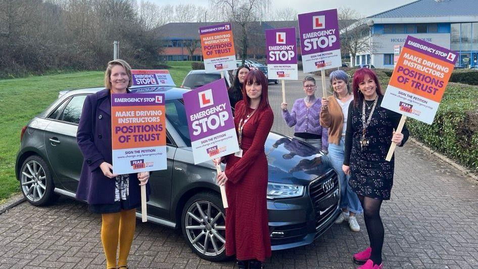 Six women stand around a grey Audi car holding placards. The placards read 'Emergency Stop' and 'Make Driving Instructors Positions of Trust'. The woman at the front left has blonde hard, a purple coat and yellow leggings. The woman in the centre has shoulder length red hair and a scarlet full length dress
