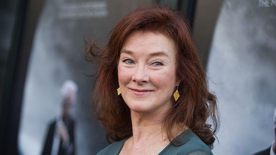 Valerie Mahaffey is pictured in front of a grey background. She has red hair and is wearing a green dress and gold earrings. She has blue eyes.