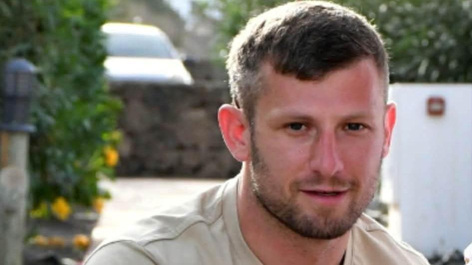 Scot James, a man with short brown hair and stubble, looking into the camera. He wears a beige top and stood in a garden.