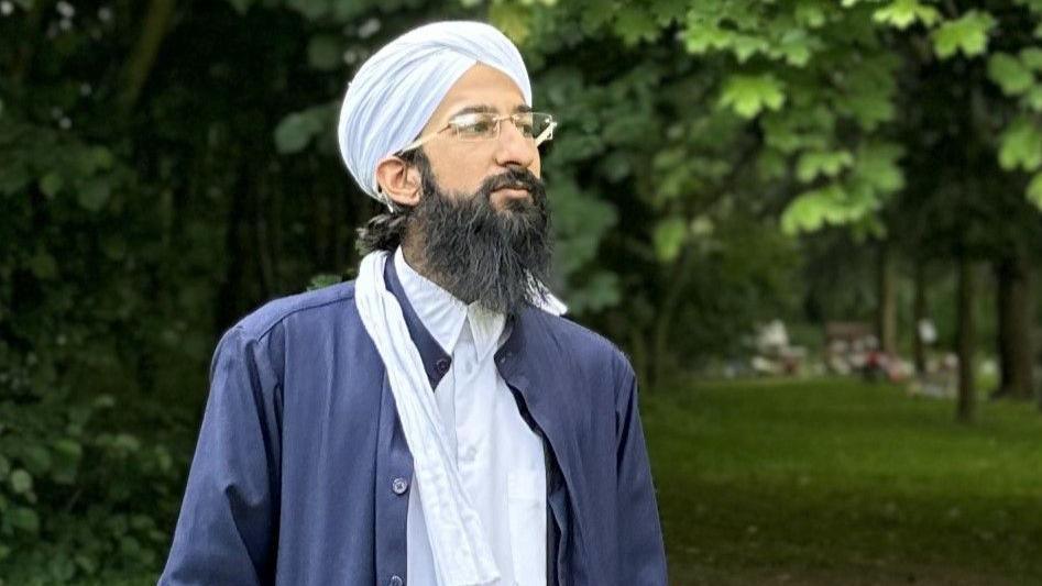 Imam Habeeb Minhas wearing a traditional turban and glasses. He has a long black beard and is wearing a blue long coat over his white clothing