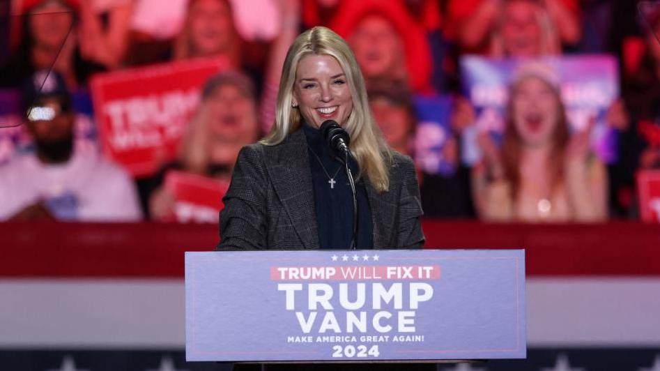 Pam Bondi addresses a crowd at a Trump rally. People are cheering and shouting behind her, holding Trump-Vance signs. Bondi has long blonde hair, and wears a grey blazer.