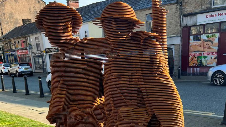 The brown steel sculpture of two soldiers walking together, one wounded, made up of slices of metal in layers to form the pair who are in Army uniforms and wearing helmets