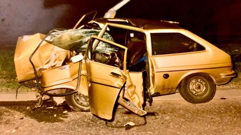 A car that has been involved in an accident. The bumper is crushed into the centre of the vehicle and the windscreen is smashed. The car which appears to be a gold in the picture's light, is crumpled on the roadside. 