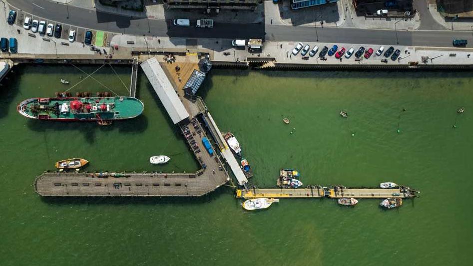 An overhead view of the Ha'penny Pier, Harwich which juts out into the port.  There are a number of boats on green looking water and cars parked along the front.