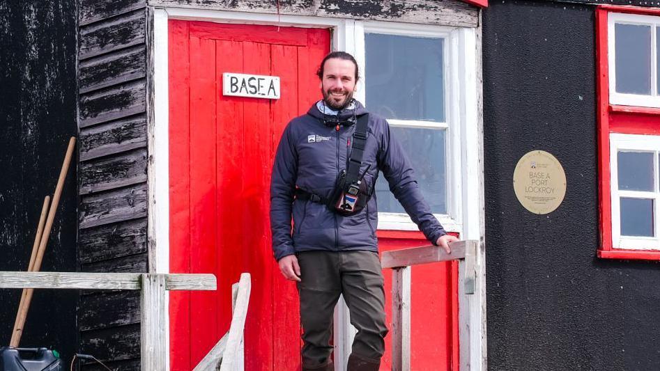 Pete Watson stands outside a wooden cabin with a red door. He is smiling, wearing a coat, trousers and scarf.