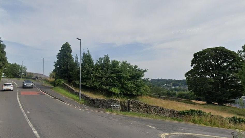 The current junction from the A591 and Thwaites Lane into the Orrest Head Farm site. There are trees and fields at the corners.