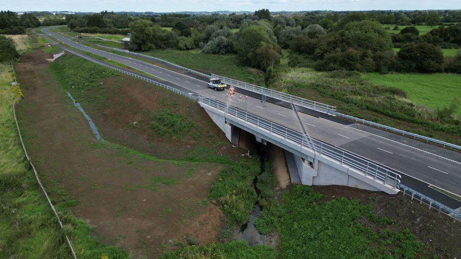 A wide shot of the road which has no cars on it apart from one maintenance vehicle with a few orange cones next to it. Part of the road is a bridge, with land sloping away underneath the road to a stream. There are lots of trees in the distance.