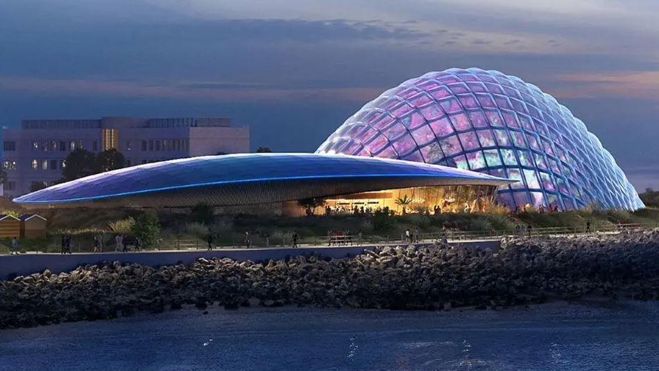 The curved roof of the proposed Eden Project Morecambe is seen in front of a glass domed, lattice framed building at the edge of a beach in an artist's drawing