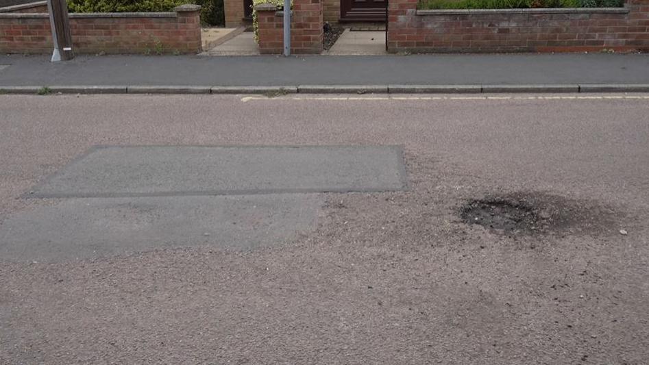 Road where large two large pothole repair patches lie net two an open pothole to the right.