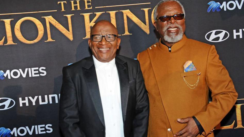 Composer Lebo M, wearing a dark suit and white shirt, and actor John Kani, wearing a tan suit on the red carpet for Lion King.