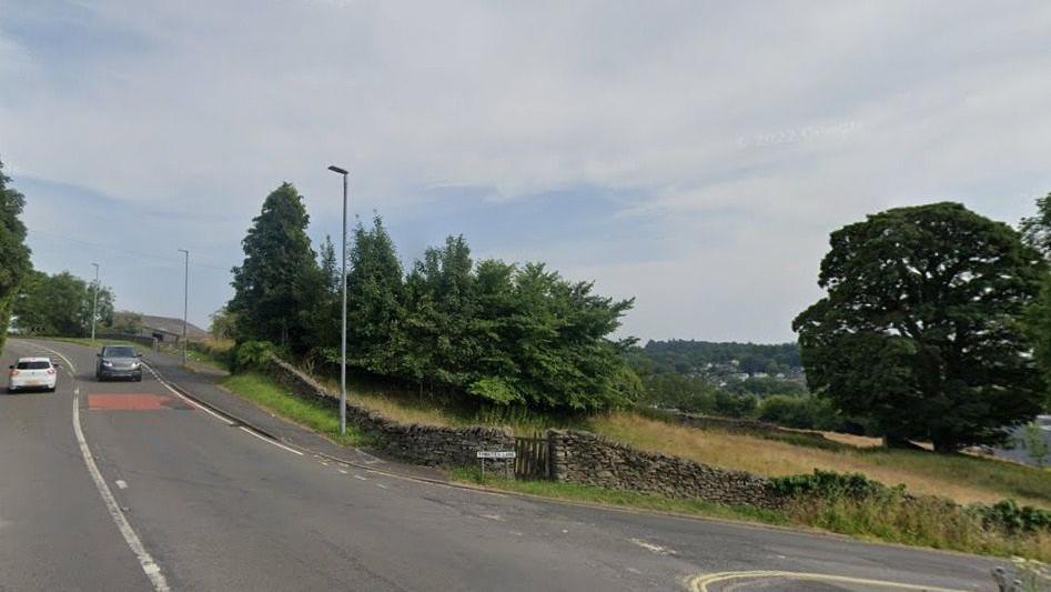 The current junction from the A591 and Thwaites Lane into the Orrest Head Farm site. There are trees and fields at the corners.