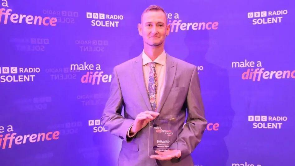 A man is standing in a suit smartly dressed, he is holding an award in his hands.  It is a BBC Make a Difference award, which has wording engraved on a glass panel.  He is standing in front of a purple background with branding for the awards on it, with the wording Make a Difference, and BBC Radio Solent.