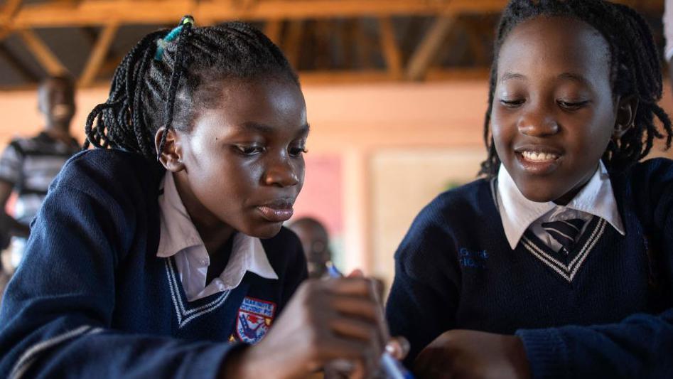 Two 13-year-old girls, who are members of the Ugandan dance group the Ghetto Kids, attend school in Kampala on 20 Apri 2023. They are looking down at a book and wearing navy school jumpers and white shirts.