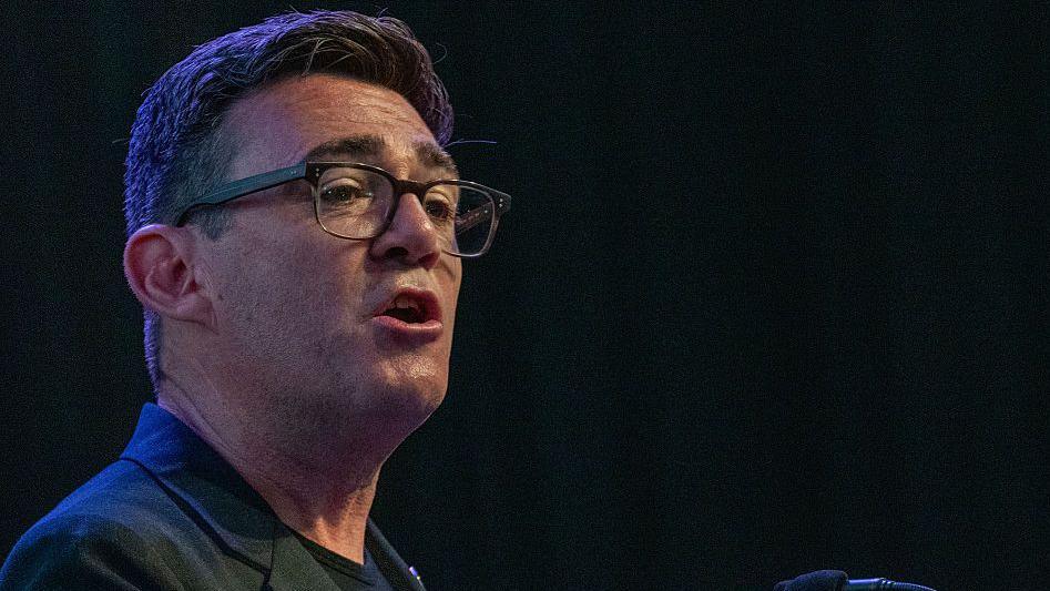 Andy Burnham wearing a black jacket with a black t-shirt talking to an audience with a black background behind him