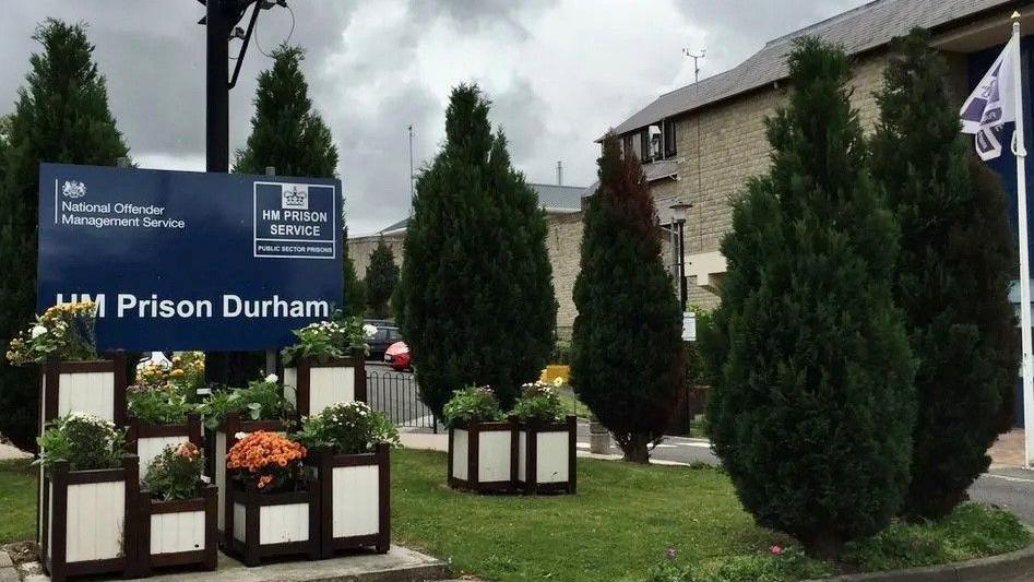 View of a sign for HMP Durham with flower boxes in front of it, and to the side a partial view of the brick structure with small trees in front of it.
